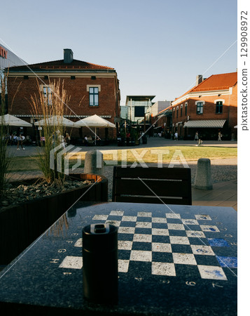 Chessboard embedded in urban outdoor table, surrounding revitalised architecture with cafes and shops, bathing in golden afternoon sunlight in Polish cityscape. Kazimierz district in Krakow, Poland Chessboard embedded in urban outdoor table, surrounding revitalised architecture with cafes and shops, bathing in golden afternoon sunlight in Polish cityscape. Kazimierz district in Krakow, Poland 129908972