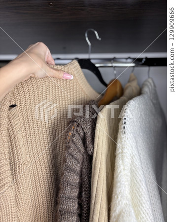 A woman sorts through clothes hanging on hangers in a closet. Reviewing and preparing seasonal wardrobe with warm knitted sweaters and jumpers for autumn winter. Minimalistic clothes organization 129909666