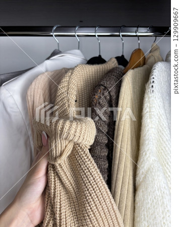 A woman sorts through clothes hanging on hangers in a closet. Reviewing and preparing seasonal wardrobe with warm knitted sweaters and jumpers for autumn winter. Minimalistic clothes organization A woman sorts through clothes hanging on hangers in a closet. Reviewing and preparing seasonal wardrobe with warm knitted sweaters and jumpers for autumn winter. Minimalistic clothes organization 129909667