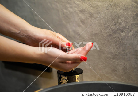 Female hands with red manicure squeeze liquid soap from soap dispenser against gray bathroom tiles. Hand washing cleaning and hygiene concept. Copy space for text 129910098