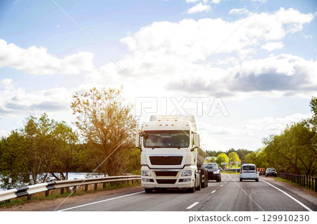 Tanker truck transports milk along country road against forest and sun in summer. Copy space for text, agriculture. Beautiful sky Tanker truck transports milk along country road against forest and sun in summer. Copy space for text, agriculture. Beautiful sky 129910230
