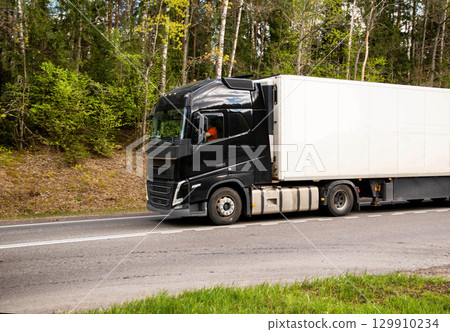 A truck with a refrigerated semi-trailer transports refrigerated cargo along a country road against the backdrop of beautiful nature and forest in summer. Copy space for text A truck with a refrigerated semi-trailer transports refrigerated cargo along a country road against the backdrop of beautiful nature and forest in summer. Copy space for text 129910234