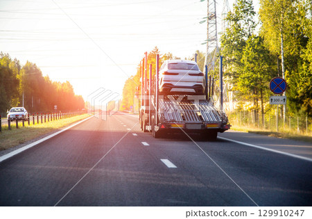 A truck with a semi-trailer for transporting cars on a country road in summer against the backdrop of a sunset. Concept international delivery of cars by car carrier. Copy space for text 129910247