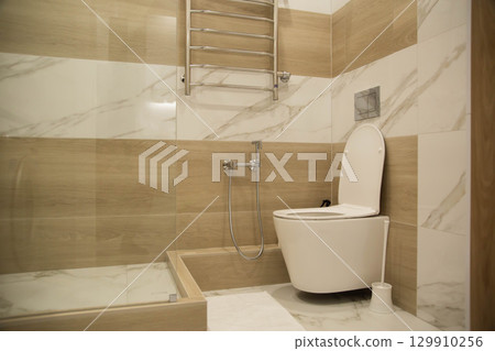 Modern wall-hung toilet against the background of a hygienic shower and porcelain tiles on the walls in a combined bathroom. Copy space for text, stylish Modern wall-hung toilet against the background of a hygienic shower and porcelain tiles on the walls in a combined bathroom. Copy space for text, stylish 129910256