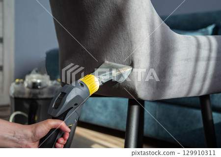 A girl s hand with a washing vacuum cleaner cleans the gray upholstery of a fabric chair in the apartment from dirt. Cleaning services, upholstered furniture cleaning A girl s hand with a washing vacuum cleaner cleans the gray upholstery of a fabric chair in the apartment from dirt. Cleaning services, upholstered furniture cleaning 129910311