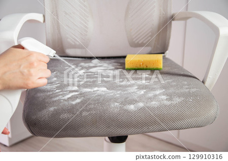 A woman's hand with a cleaning agent sprays a spray on the dirty upholstery of a computer chair. Concept of dry cleaning at home, industry A woman's hand with a cleaning agent sprays a spray on the dirty upholstery of a computer chair. Concept of dry cleaning at home, industry 129910316