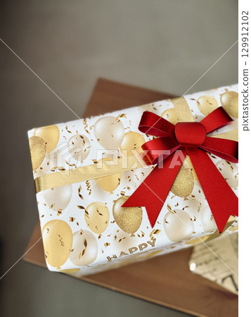 Closeup of festive gift box with gold white balloon print wrapping paper and red bow ribbon, birthday present on wooden table 129912102