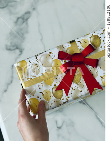 Hand holding festive birthday gift box wrapped in gold white balloon print paper with red bow ribbon on marble background Hand holding festive birthday gift box wrapped in gold white balloon print paper with red bow ribbon on marble background 129912106