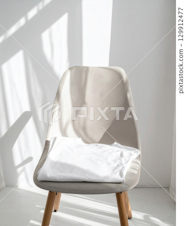 White T Shirt Neatly Folded on Light Beige Chair in a Bright Room with Shadows 129912477