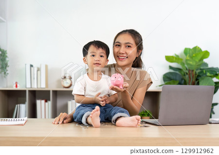 Happy mother and son showcasing piggy bank in home office 129912962