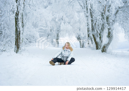 Woman sitting on a snow-covered path surrounded by snowy trees. She wears a silver jacket, smiling peacefully, enjoying the serene winter landscape. A calming winter moment 129914417