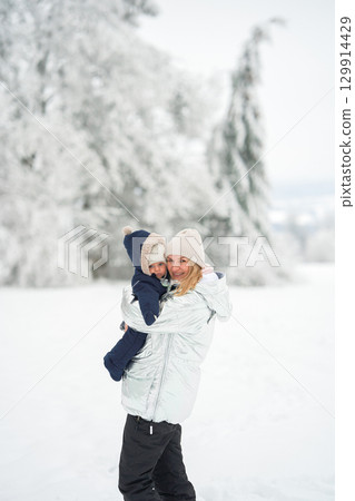 Mother and child in winter clothing enjoying a snowy day outdoors. The mother smiles warmly while holding her bundled-up child, creating a cozy and joyful winter moment 129914429