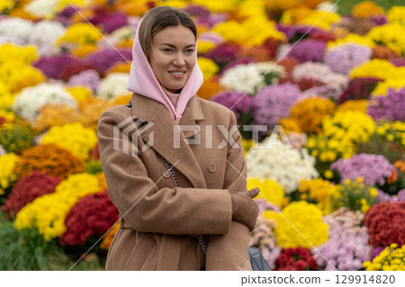 Woman in pink scarf smiles amidst vibrant floral display in a public garden during autumn afternoon 129914820