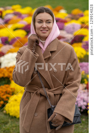 Woman in a brown coat and pink scarf smiles in a garden of colorful flowers during a sunny day 129914828