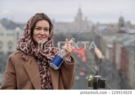 Woman in a scarf holds a bottle while enjoying a city view on a cloudy day in winter Woman in a scarf holds a bottle while enjoying a city view on a cloudy day in winter 129914829