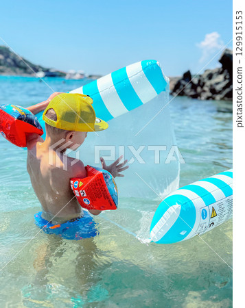 Young child enjoys a sunny day in the ocean, playing with a blue and white inflatable boat. Wearing colorful swim trunks and arm floaties, the child explores and splashes in shallow water. Young child enjoys a sunny day in the ocean, playing with a blue and white inflatable boat. Wearing colorful swim trunks and arm floaties, the child explores and splashes in shallow water. 129915153