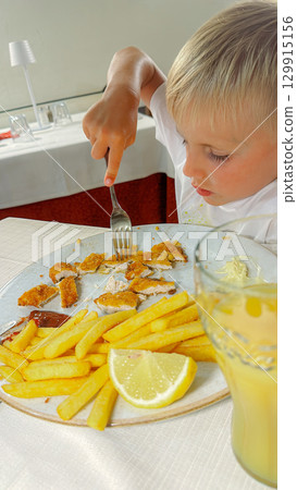 Young child intently eating breaded chicken and French fries at a restaurant. The focus is on the child's enjoyment and concentration while using a fork to interact with the meal. Young child intently eating breaded chicken and French fries at a restaurant. The focus is on the child's enjoyment and concentration while using a fork to interact with the meal. 129915156