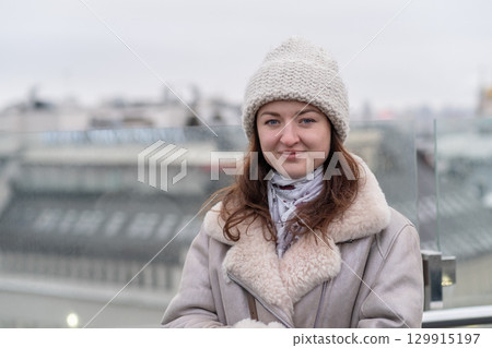 Young woman wears warm attire with winter hat while standing on rooftop in city during overcast day Young woman wears warm attire with winter hat while standing on rooftop in city during overcast day 129915197