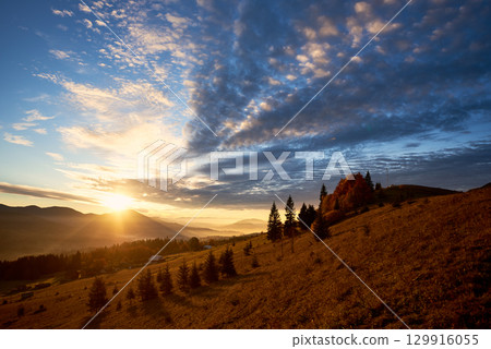 Sun rises over mountains, casting golden rays across landscape. Sky filled with dramatic clouds, creating stunning contrast with clear blue above. Warm light illuminates rolling hills and pine trees. 129916055