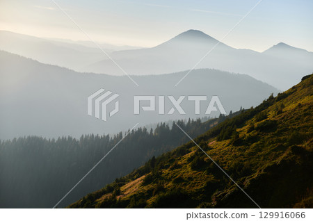 Misty morning in mountains with layers of forested hills fading into distance. Rising sun casts golden light on lush green slope, creating serene and peaceful landscape. Misty morning in mountains with layers of forested hills fading into distance. Rising sun casts golden light on lush green slope, creating serene and peaceful landscape. 129916066