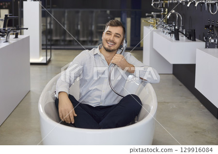 Funny sale manager helping to choose bathroom shower set sitting in bathtub, enjoy playing to bathe 129916084
