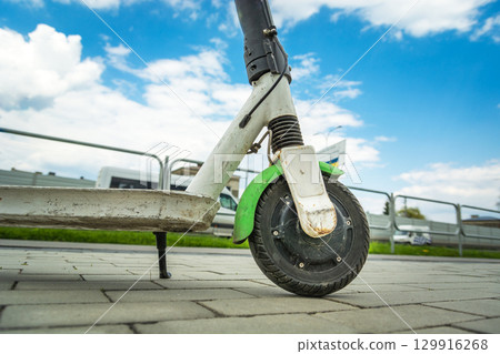 A close-up of the front of an electric scooter parked on the sidewalk next to a city street. A close-up of the front of an electric scooter parked on the sidewalk next to a city street. 129916268