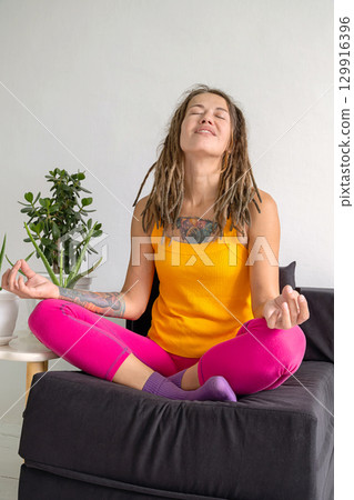 Woman with dreadlocks meditating on sofa. Practice of yoga and spiritual focus at home. 129916396