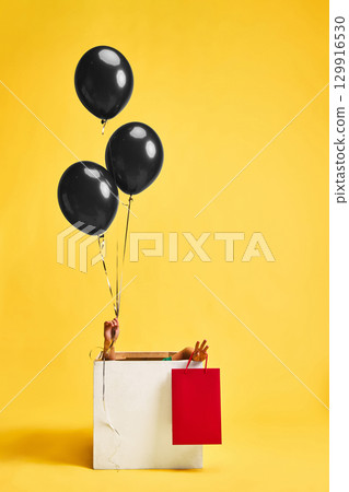 Child holding shopping bag and black balloons inside gift box on yellow background Child holding shopping bag and black balloons inside gift box on yellow background 129916530
