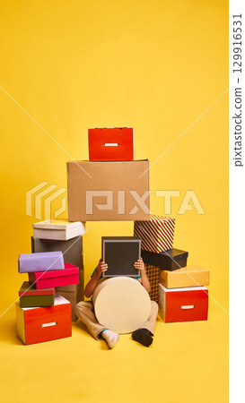 Playful child hiding behind gift boxes on bright yellow background Playful child hiding behind gift boxes on bright yellow background 129916531