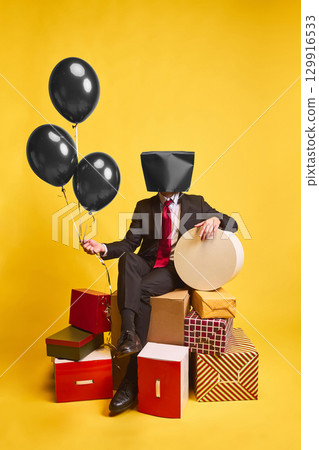 Businessman with black shopping bag on head sitting with balloons and gift boxes on yellow background Businessman with black shopping bag on head sitting with balloons and gift boxes on yellow background 129916533