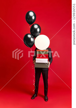 Person in suit holding stack of boxes and black balloons on red background Person in suit holding stack of boxes and black balloons on red background 129916551