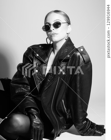 Woman black and white studio portrait with black punk leather jacket, sunglasses and without bra Woman black and white studio portrait with black punk leather jacket, sunglasses and without bra 129916944