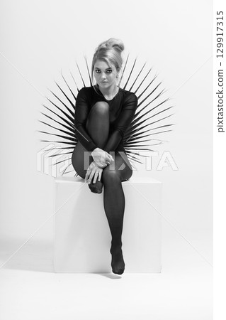 Black and white portrait of beautiful woman with tight black clothes sitting on white cube 129917315