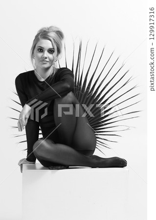 Black and white portrait of beautiful woman with tight black clothes sitting on white cube 129917316
