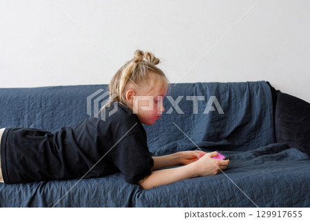 A 6 years old girl in black clothes is lying on the sofa, bored and looking at the phone gadget 129917655