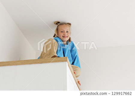 Adorable kid girl sitting on a high cabinet. Child 6 years old at home 129917662