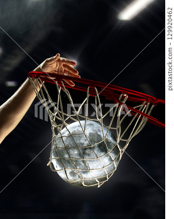 Creative composition. Basketball player dunking glowing moon through hoop in arena Creative composition. Basketball player dunking glowing moon through hoop in arena 129920462