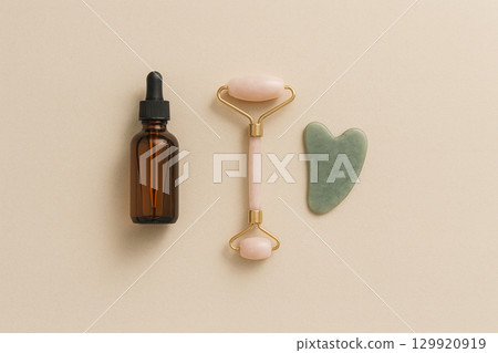 Overhead minimal composition - rose-quartz facial roller, green jade gua sha massage tool and amber glass serum dropper bottle on a neutral beige background. Clean, unbranded self-care concept. Overhead minimal composition - rose-quartz facial roller, green jade gua sha massage tool and amber glass serum dropper bottle on a neutral beige background. Clean, unbranded self-care concept. 129920919