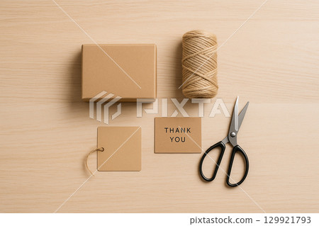 Top-down flat lay - kraft cardboard box, ball of twine, metal scissors, blank hang tag and THANK YOU card on a light wooden desk. Neutral, unbranded packaging shipping concept with copy space. 129921793