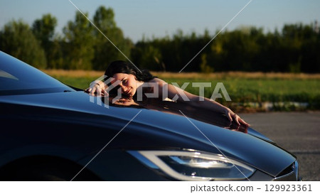 Beautiful smiling girl bought new electrical vehicle and hugging hood auto. Happy young woman enjoying modern electric automobile. Concept of buy or rent car. Slow motion 129923361