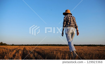 Adult agronomist in straw hat walking among barley plantation at dusk. Female farmer going through wheat meadow at sunset. Beautiful scenic landscape. Concept of agricultural business. Slow motion 129923368