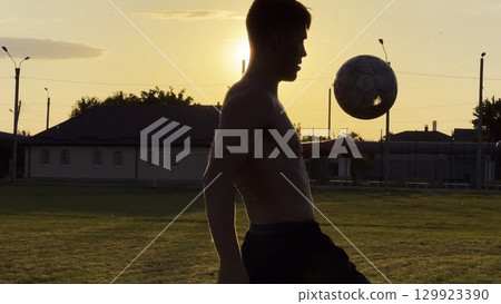 Young man juggling soccer ball on stadium at sunset. Professional footballer kicking ball at green field. Sportsman practicing tricks at meadow with sunlight at background. Freestyle football 129923390