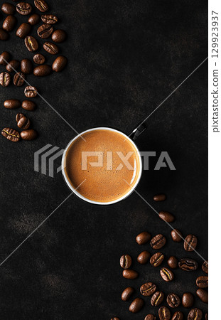 A cup of coffee sits on a dark, textured background, surrounded by scattered roasted coffee beans. The image is shot from a top-down perspective. 129923937