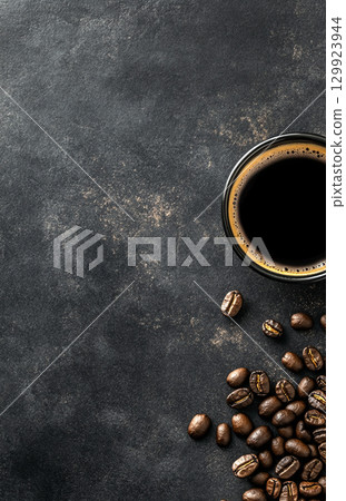 A cup of coffee sits on a dark, textured background, surrounded by scattered roasted coffee beans. The image is shot from a top-down perspective. 129923944