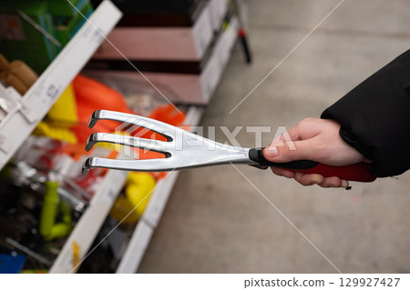 Hand holding a three-pronged garden cultivator tool in a retail store aisles showcasing gardening supplies Hand holding a three-pronged garden cultivator tool in a retail store aisles showcasing gardening supplies 129927427