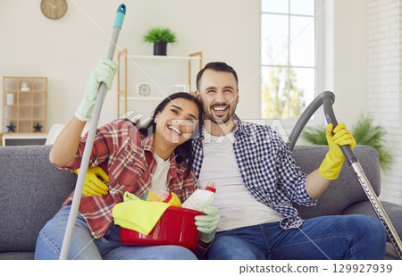 Happy couple with household cleaning supplies in hands sitting on sofa at home 129927939
