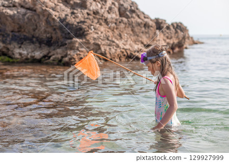 Young girl exploring shallow water with net, wearing snorkeling gear, surrounded by rocky shoreline Young girl exploring shallow water with net, wearing snorkeling gear, surrounded by rocky shoreline 129927999