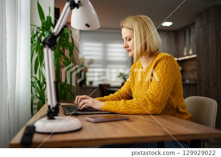 Focused woman in yellow sweater working on laptop at home desk with bright lamp and plants in a cozy atmosphere 129929022