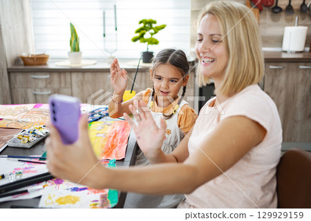 Mother and daughter engaged in a cheerful video call while painting together at home in a cozy kitchen setting Mother and daughter engaged in a cheerful video call while painting together at home in a cozy kitchen setting 129929159