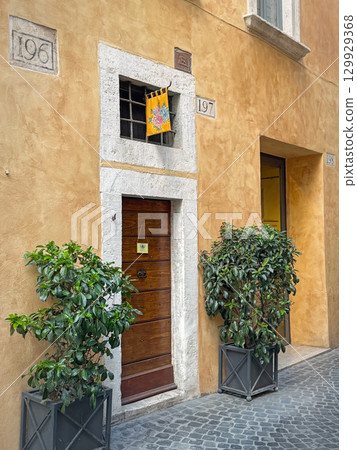 ROME, ITALY - July 30, 2025 Narrow street view showing an old yellow building facade with a wooden door and a colorful flag hangs above. Two potted green plants flank the entrance 129929368